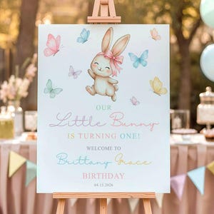 May include: A white sign on a wooden easel with a watercolor illustration of a bunny and butterflies. The sign reads "Our Little Bunny Is Turning One!" and welcomes guests to Brittany Grace's birthday on 04.15.2026. Pastel colors are used.