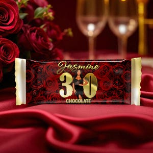 Pode incluir: Uma barra de chocolate com um fundo de padrão de rosas vermelhas. A barra apresenta o nome "Jasmine" em letras douradas, o número "30" em ouro e a palavra "CHOCOLATE". Uma mulher de vestido preto também está presente.