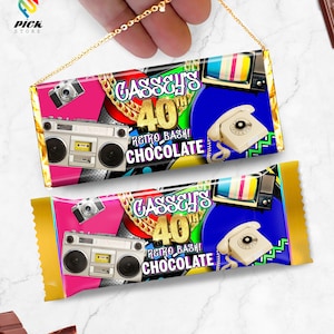 以下が含まれることがあります： レトロなテーマのチョコレートバーの包装が2つ。包装はピンク、ブルー、ゴールドで、ブームボックス、電話、カメラ、テレビの画像が描かれています。包装のテキストには「Cassey's 40th Retro Bash! Chocolate」と書いてあります。