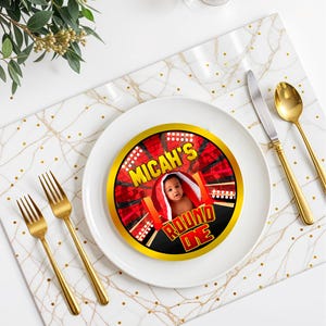 May include: Round one birthday party plate with a gold border and a red and black boxing ring design. The plate features a photo of a baby boy in a red robe with the name Micah and the text "Round One".