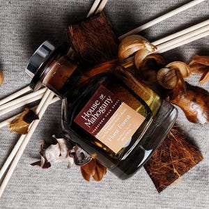 Może przedstawiać: Brązowa szklana butelka ze srebrną zakrętką i etykietą z napisem "House of Mahogany Transform Your Space" i "Hurst Forest" na brązowym tle. Butelka jest otoczona drewnianymi patyczkami i elementami dekoracyjnymi.