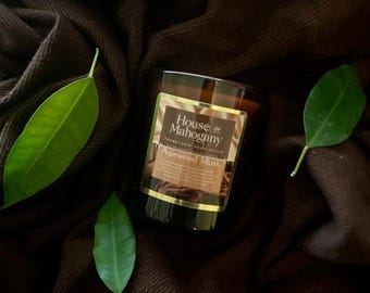 Może przedstawiać: Szklany słoik na świecę w kolorze brązowym z  złotą etykietą z napisem "House of Mahogany Transform Your Space Agarwood Musk". Świeca stoi na brązowej powierzchni z tkaniny z zielonymi liśćmi.