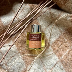 Puede incluir: Una botella de vidrio transparente con una tapa dorada y una etiqueta marrón. La etiqueta dice "House of Mahogany" y "Hurst Forest". La botella está sobre una tela de punto marrón y blanca.