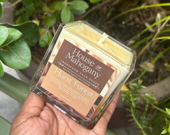 May include: A square glass candle jar with a yellow candle inside. The label reads "House of Mahogany Transform Your Space From The Signature Scent Collection Hurst Forest".