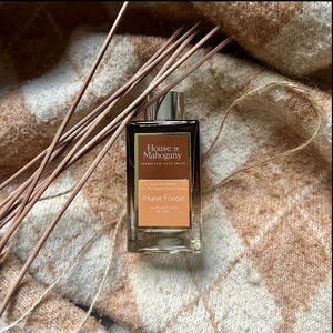 Może przedstawiać: Szklana butelka z luksusowym dyfuzorem trzcinowym Hurst Forest z kolekcji The Sophisticated Scents Collection marki House of Mahogany. Butelka jest brązowa z złotą etykietą i leży na brązowo-białym dzianym kocu.