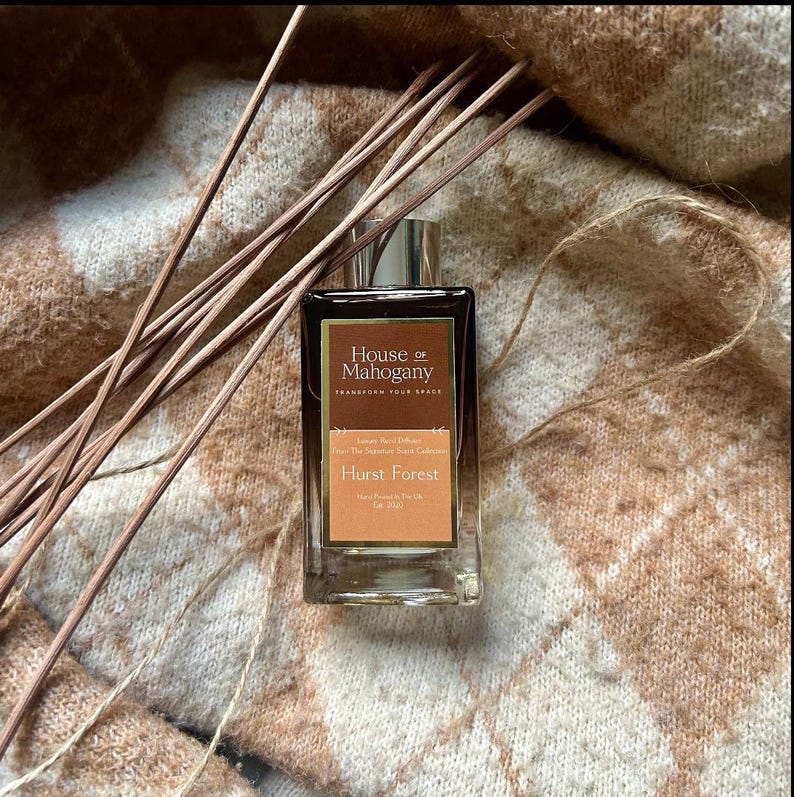 Może przedstawiać: Szklana butelka z luksusowym dyfuzorem trzcinowym Hurst Forest z kolekcji The Sophisticated Scents Collection marki House of Mahogany. Butelka jest brązowa z złotą etykietą i leży na brązowo-białym dzianym kocu.