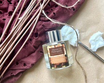 Może przedstawiać: Przezroczysta szklana butelka ze srebrną zakrętką i etykietą z napisem "House of Mahogany" i "Agarwood Musk". Butelka stoi na brązowej powierzchni z kawałkiem sznurka i drewnianymi patyczkami.