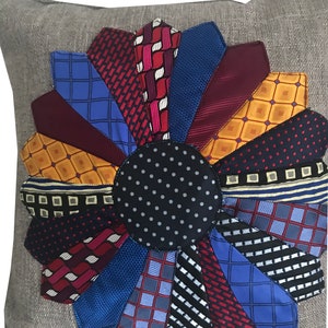 May include: A decorative cushion featuring a large flower design crafted from upcycled neckties. The flower is composed of various colours and patterns of neckties, including blue, red, yellow, and black. The cushion is made from a light brown fabric.