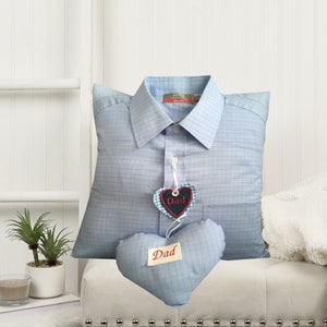 May include: A blue plaid shirt cushion with a heart-shaped tag that says "Dad". Another heart-shaped cushion with the word "Dad" is also present. The cushion is on a white ottoman with silver studs. A small plant and candle are on a side table.