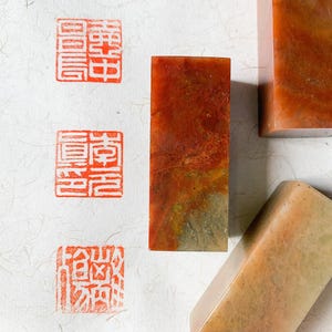 May include: Close-up of several rectangular stone seals with carved Chinese characters and red ink stamps on textured paper. The seals are various shades of orange, brown, and beige. The paper has a subtle, natural pattern.