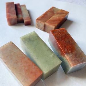 May include: Several rectangular stone seals in shades of red, green, and tan are displayed. The seals are arranged on a white surface, highlighting their polished surfaces and natural colour variations. Each seal is a unique piece.