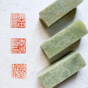 May include: Three rectangular, light green jade seals with rounded edges are arranged diagonally. Three red Chinese characters are stamped on the left side of the image, showcasing the seals' potential use. The seals are on a textured white surface.
