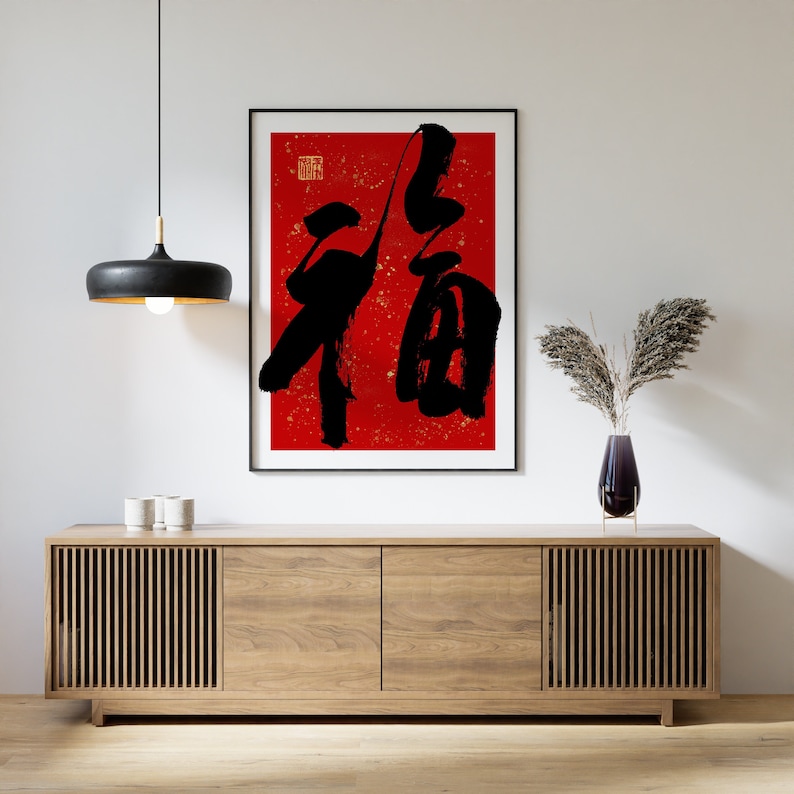 May include: A black and white calligraphy print of the Chinese character "福" (f&uacute;), meaning "good fortune" or "happiness", on a red background with gold flecks. The print is framed in black and hanging on a white wall above a wooden cabinet.