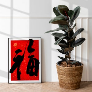 May include: A black and white calligraphy print of the Chinese character "福" (f&uacute;), meaning "good fortune" or "happiness", on a red background. The print is framed in black and displayed on a white wall next to a potted rubber plant.