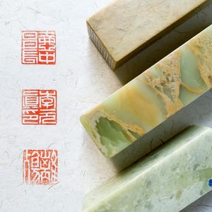 May include: Three rectangular stone seals with smooth surfaces. One is tan, one is pale green with yellow veining, and one is light green. To the left, three red ink stamps are imprinted on textured white paper, each featuring intricate Chinese characters.