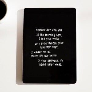 Op de afbeelding: Een zwarte tablet toont witte tekst: "Another day with you...In your embrace, my heart takes wings." Een witte koffiekop met zwarte koffie staat in de linkerbovenhoek. De achtergrond is wit.