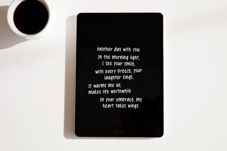Op de afbeelding: Een zwarte tablet toont witte tekst: "Another day with you...In your embrace, my heart takes wings." Een witte koffiekop met zwarte koffie staat in de linkerbovenhoek. De achtergrond is wit.