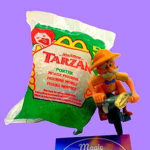 May include: A plastic bag with a McDonald's Happy Meal toy inside. The toy is a yellow and orange figure of Porter from the Disney movie Tarzan. Porter is riding a motorcycle. The bag has the words "Tarzan" and "Porter" printed on it.
