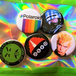 Puede incluir: Cinco pines de botón coloridos con varios diseños. Los pines presentan una cara sonriente verde con el texto "Mr. Yuk Means No! Stay Away!" un pin blanco y negro con un diseño rojo y blanco y el texto "Devo", un pin blanco y negro con una cabeza de robot, un pin blanco y negro con la cara de una persona y un pin arcoíris con el texto "Polaroid".