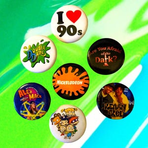 May include: Nine round button badges featuring various Nickelodeon shows and logos from the 1990s. The badges feature the logos for "Double Dare", "Are You Afraid of the Dark?", "Nickelodeon", "Alex Mack", "Rugrats", and "Kenan & Kel". One badge features a heart with the text "I 90s".
