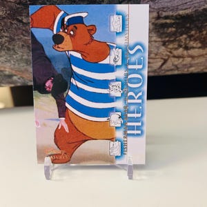 May include: A trading card featuring a cartoon bear in a sailor outfit with blue and white stripes. The card has the word "HEROES" in blue and white text. The card is propped up on a clear plastic stand.