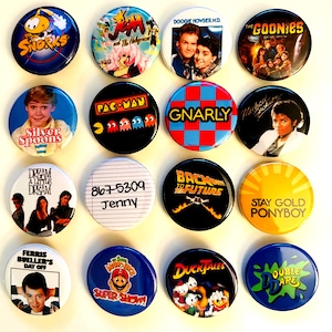 May include: A selection of vibrant, circular pin badges showcasing pop culture icons. Designs include characters and logos from "The Snorks," "Pac-Man," "Doogie Howser, M.D.," "The Goonies," "Silver Spoons," "Back to the Future," and more. Each badge is approximately 2.5 cm in diameter.