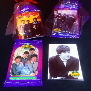 Könnte beinhalten: Zwei verpackte Pakete und eine violette Folienverpackung mit dem Branding The Beatles Collection und Bildern der Bandmitglieder. Eine separate rechteckige Karte zeigt ein Schwarz-Weiß-Porträt von Paul McCartney mit dem Logo der Kollektion.