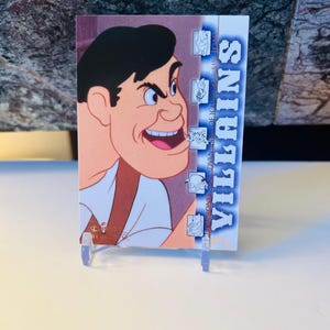 May include: A trading card featuring a cartoon illustration of a man with a sinister grin. The card has the text "VILLAINS" in a vertical blue font. The card is propped up on a clear plastic stand.