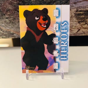 Könnte beinhalten: Eine Sammelkarte mit einer Cartoon-Bären-Illustration. Der Bär ist schwarz mit roter Nase und Mund und lächelt. Die Karte trägt das Wort "HEROES" in blauer Schrift und kleine Charakterillustrationen.