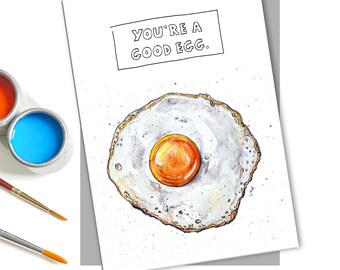 May include: A white card with the text "YOU'RE A GOOD EGG." in black lettering. A watercolor illustration of a fried egg with a bright orange yolk is below the text.