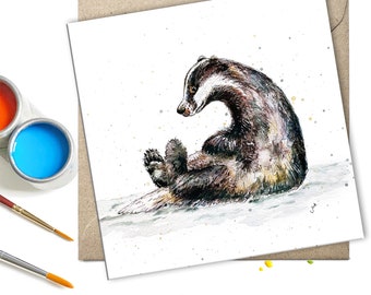 May include: A watercolor painting of a badger sitting on its haunches with its paws tucked in. The badger is brown and black with a white stripe down its face. The background is white with a few specks of gold paint.