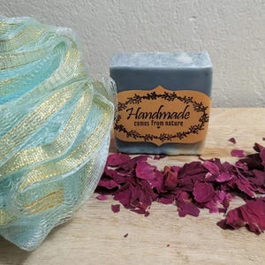 Peut inclure: Un pain de savon artisanal avec une étiquette indiquant "Handmade comes from nature" est posé à côté d'une éponge de bain en filet bleu clair avec des accents dorés et d'une pile de pétales de rose séchés. Le savon est gris et blanc.