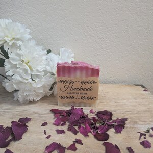 Peut inclure: Un savon artisanal avec un design en couches rose et blanc. Le savon porte une étiquette indiquant "Handmade come from nature". Des pétales de rose séchés sont dispersés autour du savon. Des fleurs blanches sont en arrière-plan.
