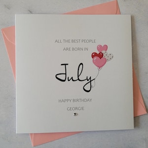 May include: A white card with a pink envelope. The card has the text "All the best people are born in July" with a black script font. There are three pink and red heart-shaped balloons with white polka dots. The text "Happy Birthday Georgie" is at the bottom of the card.