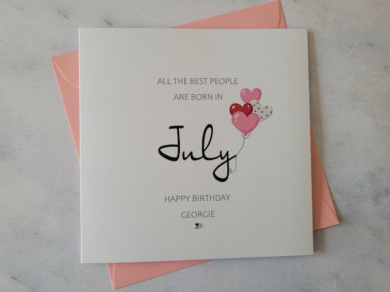 May include: A white card with a pink envelope. The card has the text "All the best people are born in July" with a black script font. There are three pink and red heart-shaped balloons with white polka dots. The text "Happy Birthday Georgie" is at the bottom of the card.