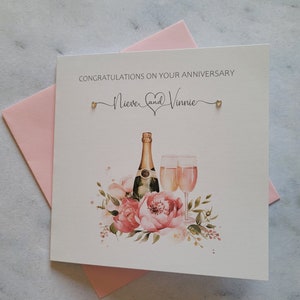 May include: A white card with the text "Congratulations on your Anniversary" and the names "Nieve and Vinnie" with a heart symbol between them. The card features a watercolour illustration of a bottle of champagne and two champagne flutes with pink flowers.