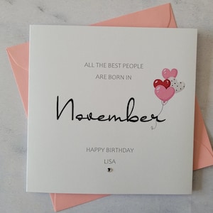 Pu&ograve; includere: Un biglietto bianco con il testo "All the best people are born in November" e un disegno di palloncini a forma di cuore rosa e rosso. Il biglietto dice anche "Happy Birthday Lisa".