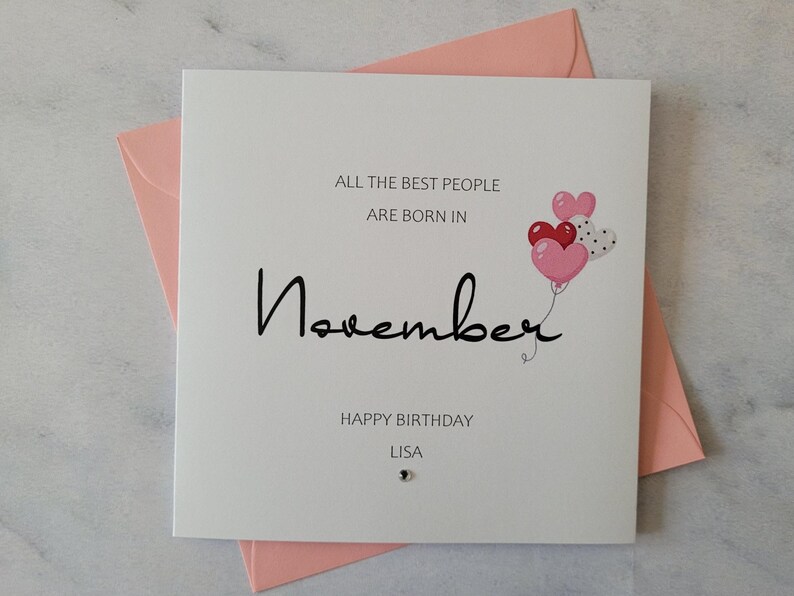 Pu&ograve; includere: Un biglietto bianco con il testo "All the best people are born in November" e un disegno di palloncini a forma di cuore rosa e rosso. Il biglietto dice anche "Happy Birthday Lisa".