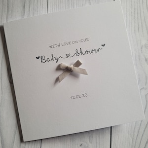 May include: A white card with the text "With love on your Baby Shower" and a small, light gray bow. The date "12.02.23" is printed below the text.