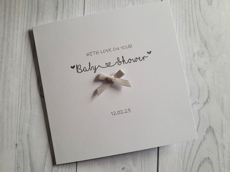 May include: A white card with the text "With love on your Baby Shower" and a small, light gray bow. The date "12.02.23" is printed below the text.