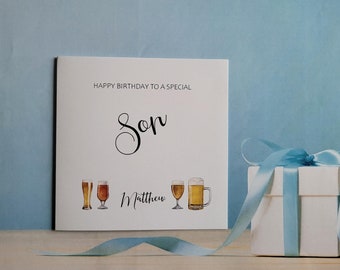 May include: A white card with the text "Happy Birthday to a Special Son Matthew" and a drawing of four beer glasses. The card is sitting on a wooden surface with a blue ribbon and a white gift box.