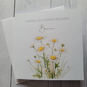 May include: A white card with a watercolour illustration of white daisies with yellow centres. The card says "Wishing you a speedy recovery Brian".