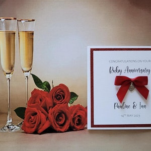 May include: A white card with red glitter trim and a red bow with a silver heart charm. The card reads "Congratulations on your Ruby Anniversary" and "Pauline & Ian 14th May 2023". Two champagne flutes and a bouquet of red roses are in the background.