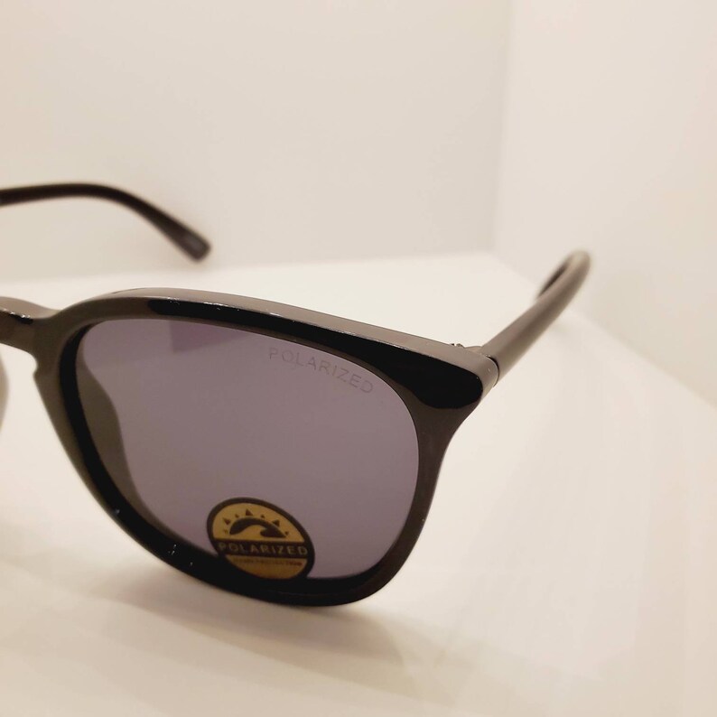 May include: Black sunglasses with polarized lenses. The lenses are tinted a dark grey color. The sunglasses have a black frame and the words "POLARIZED" are printed on the right lens.