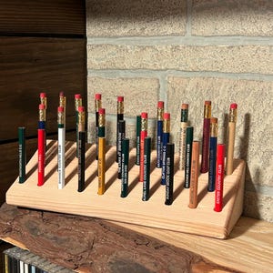 May include: A collection of pencils with red erasers, each displaying the name of a golf club. The pencils are arranged in a wooden holder, showcasing various club names like "Wentworth Hills G.C." and "Swansea Country Club". The pencils are displayed on a wooden shelf.