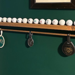 May include: A wooden golf ball display shelf with a row of golf balls and three hanging tags. The shelf is mounted on a dark green wall. The golf balls are white with various logos.