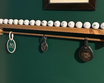 May include: A wooden golf ball display shelf with a row of golf balls and three hanging tags. The shelf is mounted on a dark green wall. The golf balls are white with various logos.