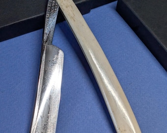 May include: A vintage straight razor with a cream-colored handle and a silver blade. The blade has etched details and text. The razor is open, displaying the sharp edge. Two black boxes are in the background, suggesting a case.