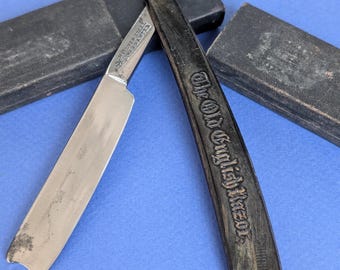 May include: An open straight razor with a dark handle, engraved with "The Old English Razor." The silver blade is partially open, with text visible on the blade. The razor is next to a black case on a blue surface.