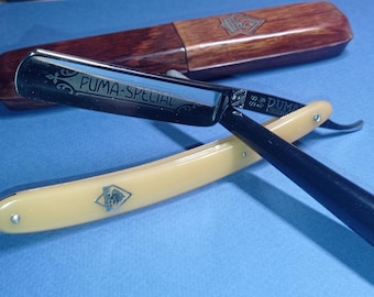 Puede incluir: Una navaja de afeitar con mango color crema y hoja negra, marcada "PUMA-SPECIAL". La navaja está abierta, apoyada sobre una superficie azul, con una funda de madera en el fondo. La funda tiene un logotipo.
